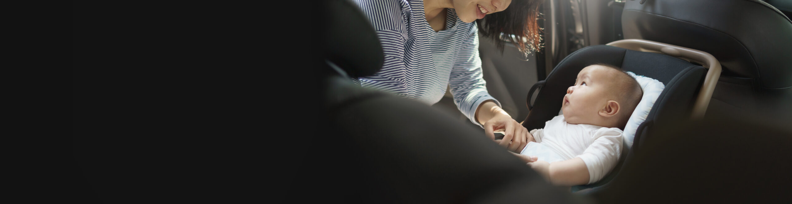 A smiling person with dark hair, wearing a striped shirt, is leaning over a baby in a car seat. The baby, dressed in white, is looking up at the person, both appearing cheerful. The background is mostly dark, highlighting the interaction between the two.