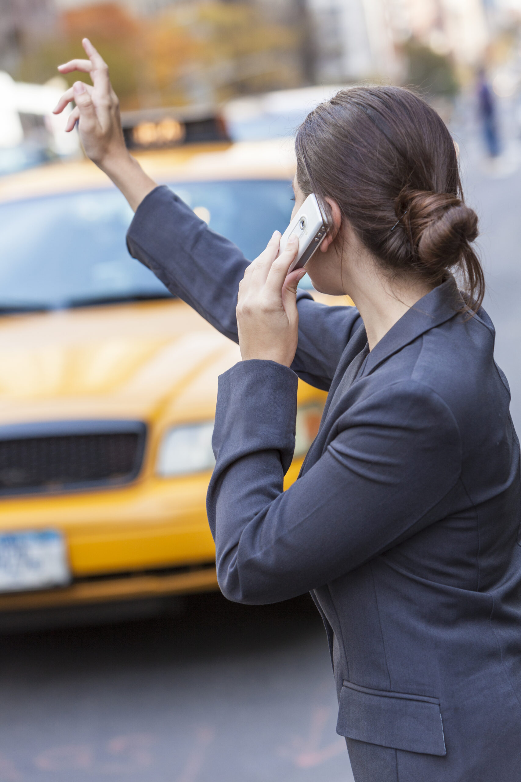 A woman wearing a dark blazer is standing on the street, talking on a cellphone with her right hand, and raising her left arm to hail a yellow taxi cab that is approaching. The background is slightly blurred, showing other vehicles and buildings.