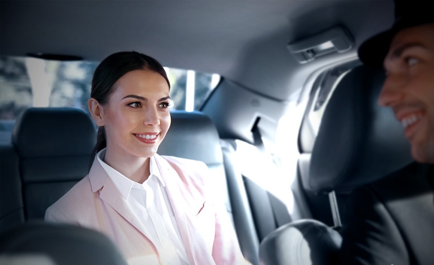 A woman in a pink blazer and white shirt smiles while sitting in the back seat of a taxi. A taxi driver, whose face is partially visible and who is wearing a hat, appears to be speaking to her from the front seat. Sunlight is streaming through the car's windows.