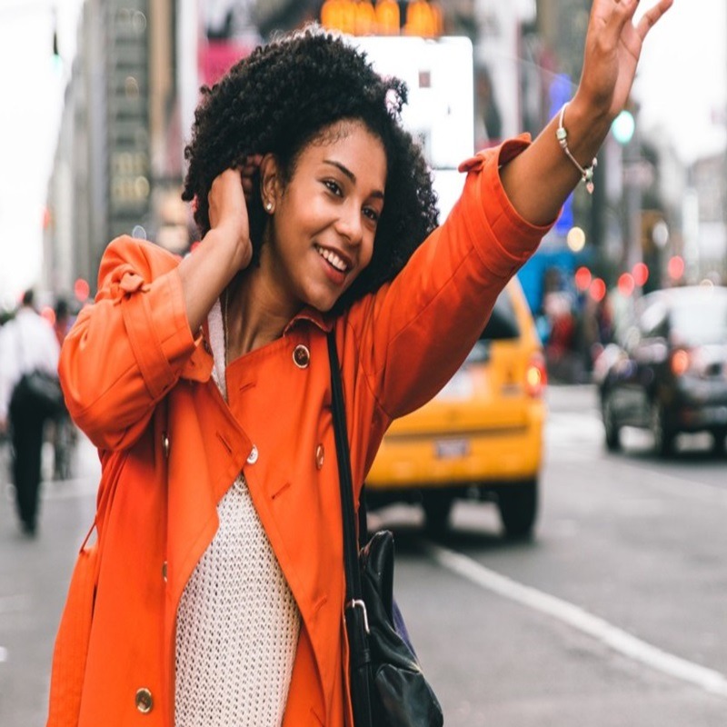 A woman with curly hair, wearing an orange coat and white top, is smiling and waving her hand, seemingly hailing a taxi, on a busy city street. A yellow taxi cab and other pedestrians are visible in the background.